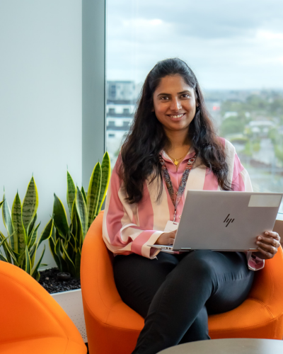 Anafrid is sitting in an orange chair, in front of a window, holding an open laptop and smiling at the camera. 