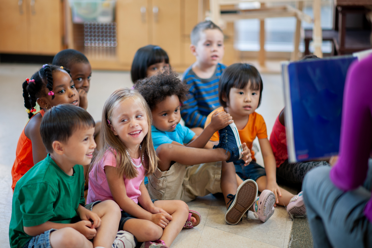 A female daycare teacher sits in a chair in front of a large group of students as she reads them a story. They students are all dressed casually and sitting with their legs crossed as they smile and listen attentively.