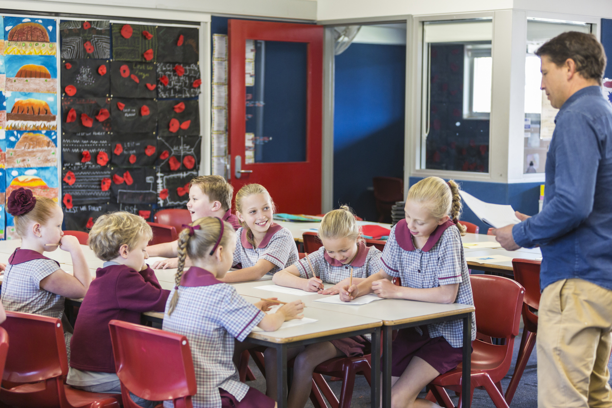 School children working with their teacher in the classroom. The kids are on one big table and the teacher is standning up on the right. 