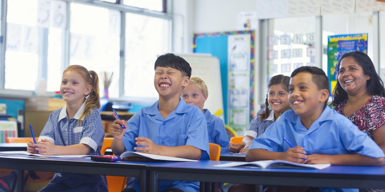 Primary school students. They are happy and smiling. They are wearing their school uniform in the classroom. A teacher is sitting with them while they are watching something. Australia