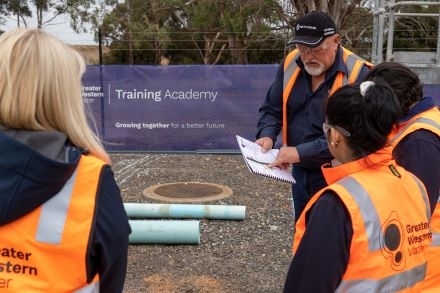 Four workers in high-visibility vests at a Greater Western Water Training Academy site, listening to one person holding a clipboard and discussing notes.