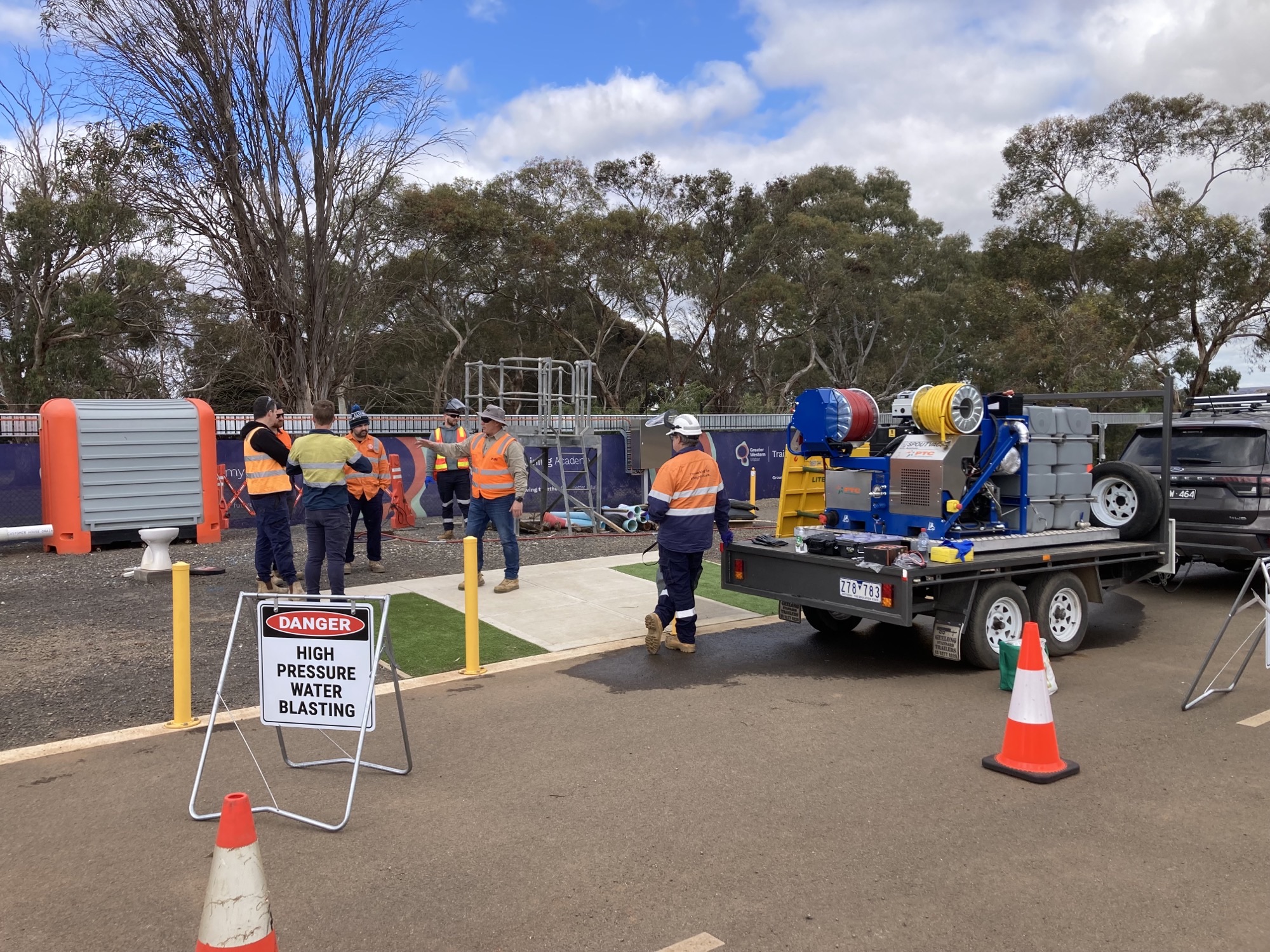 Workers in high-visibility vests gather near equipment at an industrial site marked with signs indicating "Danger High Pressure Water Blasting".