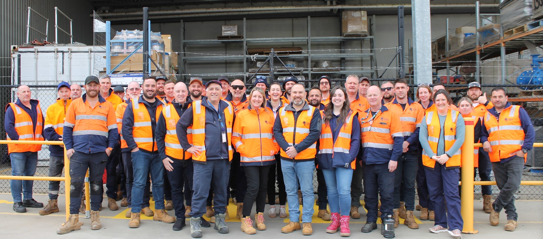 A group of people wearing high-visibility clothing stands in front of a warehouse. They are smiling and posing for a group photo. The background shows shelves with packed items. The scene appears to be at an industrial site.