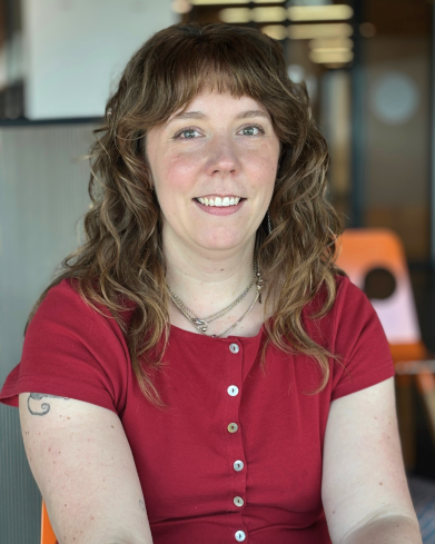 Rachel is sitting and smiling at the camera in an indoor environment and is wearing a red top