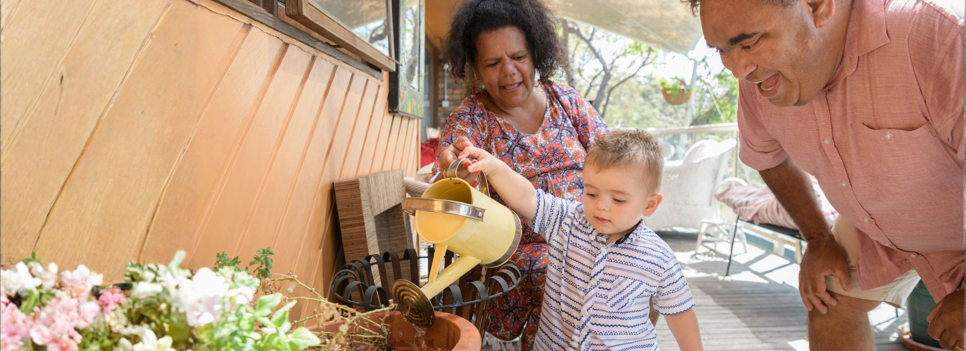 A small child with a watering can receiving help from two adults to water a plant.