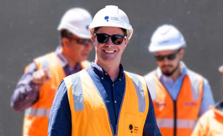 Warren is wearing a bright orange protective vest and a white helmet witht he GWW log. Two other workers are in the background dressed similarly.