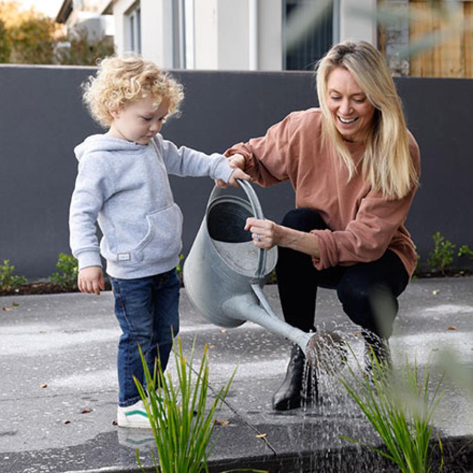 A mother and child using a watering can to water some plants in their driveway.