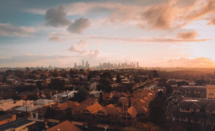 A rooftop view of Melbourne beneath a golden sky. Houses line the streets and in the distance the tall buildings of Melbourne's CBD can be seen.
