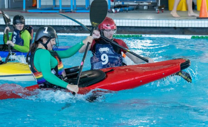 Action shot of young people in a pool using kayaks to play a game of polo. Their paddles are in the air and the water in splashing.