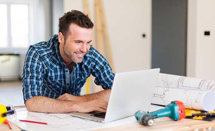 A tradesman is sitting at a desk looking at a laptop computer, the desk also holds property plans an electric drill and several other tools of trade.