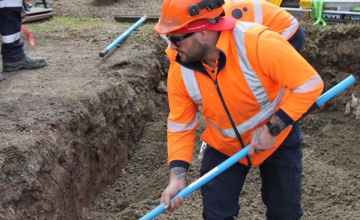 A construction worker in a high visibility orange jacket and helmet uses a shovel at a dig site with heavy machinery in the background.