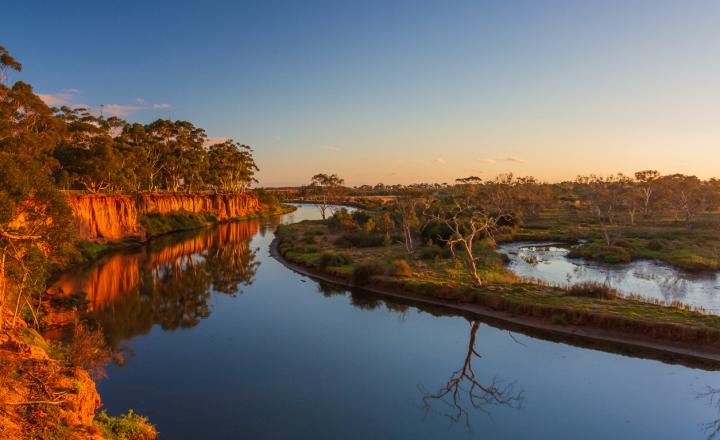 The Werribee River, photograph taken from the bank so showing the deep blue colour of the water and the sun highlighting the red rocks making up the bank of the river.