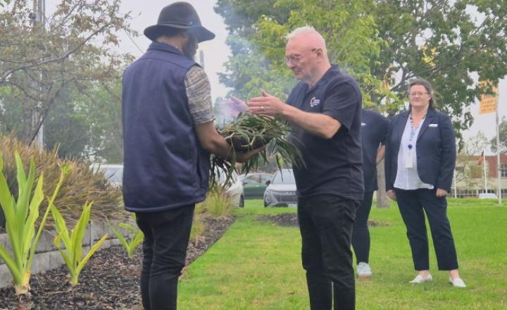 A person in a hat holds a smoking bundle of leaves, offering it to another individual in a dark shirt standing on a grassy area. Two people stand in the background watching the interaction. Trees and banners are in the background.