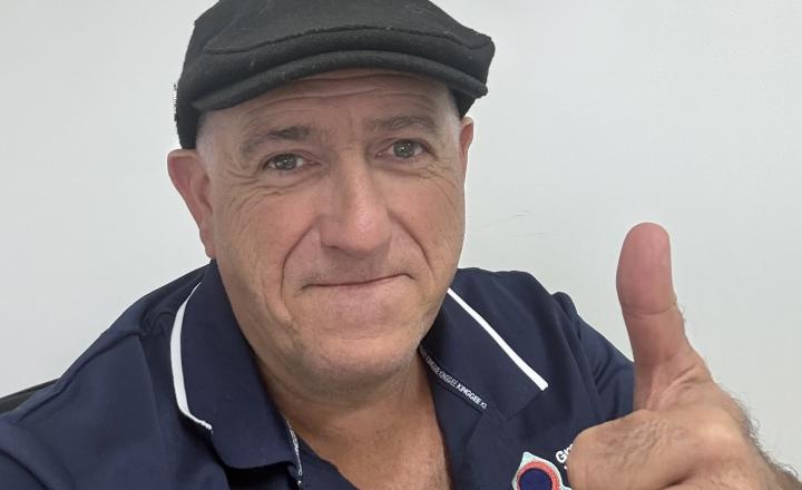 Headshot of Richard wearing a dark flat cap and a navy-blue shirt gives a thumbs-up while sitting indoors. The shirt has the GWW logo near the collar.