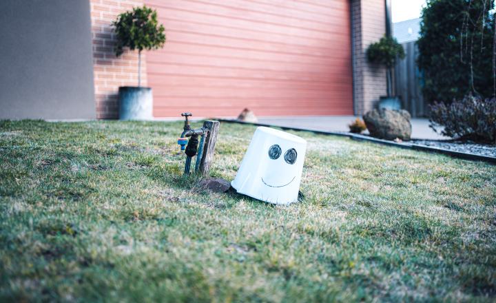 Bucket with a drawn smiling face tipped on a grassy lawn beside a garden tap, in front of a suburban house with a brick wall and closed garage door.