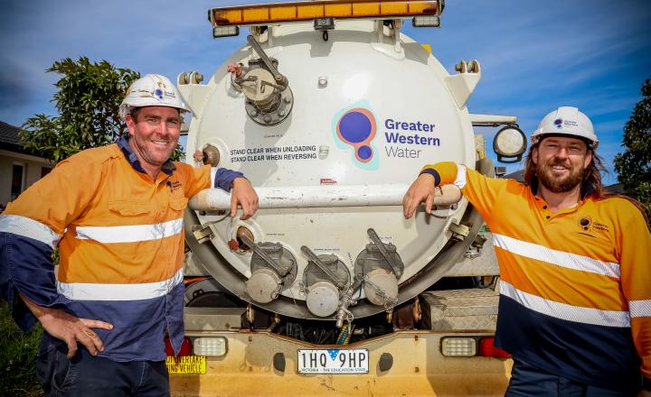 Two Greater Western Water crew members wearing high-visibility orange and navy work uniforms and white hard hats stand on either side of a large white service truck. The rear tank of the truck displays the Greater Western Water logo and safety signage. The workers rest their arms on a metal rail at the back of the vehicle, with residential houses, trees and a bright blue sky visible in the background.