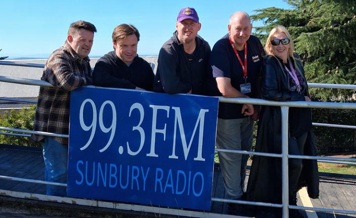 Members of the Sunbury Radio community standing in front of the station's banner, saying "99.3FM Sunbury Radio"