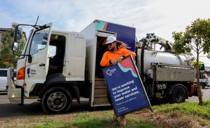 Man in protective clothing is standing next to a water tanker and putting a sign out on the naturestrip
