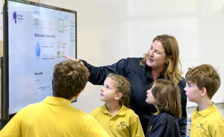 A classroom group gathers around a large interactive display showing school water-use data. One adult points to information on the screen while four primary school students in yellow school uniforms look on. The display includes graphs, water consumption figures and rainfall information as part of a water conservation or sustainability activity.
