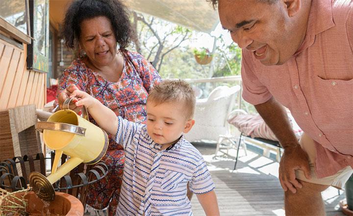 A small child with a watering can receiving help from two adults to water a plant.