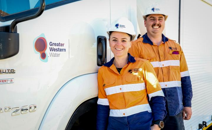 Two Greater Western Water field workers wearing high-visibility orange and navy uniforms and white safety helmets stand beside a white service truck displaying the Greater Western Water logo. The workers are positioned next to the truck door, with the vehicle filling most of the background in a bright outdoor setting.