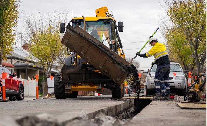Photo of a digger in an suburban street with a person using a shovel to disperse gravel from the machine