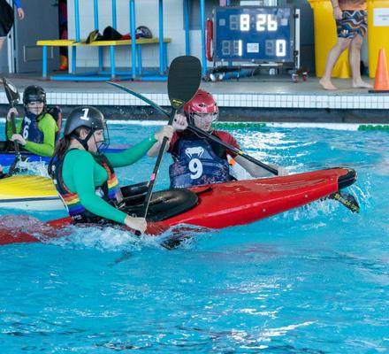 Action shot of young people in a pool using kayaks to play a game of polo. Their paddles are in the air and the water in splashing.