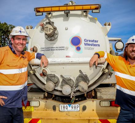 Two Greater Western Water crew members wearing high-visibility orange and navy work uniforms and white hard hats stand on either side of a large white service truck. The rear tank of the truck displays the Greater Western Water logo and safety signage. The workers rest their arms on a metal rail at the back of the vehicle, with residential houses, trees and a bright blue sky visible in the background.