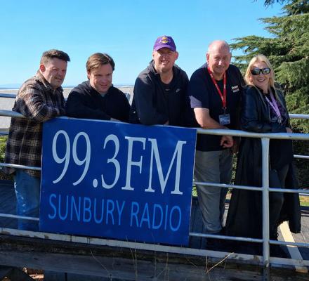Members of the Sunbury Radio community standing in front of the station's banner, saying "99.3FM Sunbury Radio"