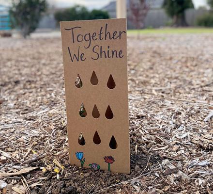 A photograph of a card, it is coloured brown and made of thin cardboard. The cover says "Together we shine" beneath this is three rows of rain drops cut out of the cardboard and at the bottom three primitive drawings of small trees.