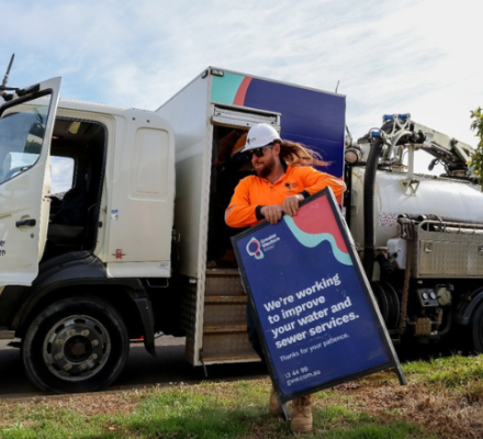 Man in protective clothing is standing next to a water tanker and putting a sign out on the naturestrip