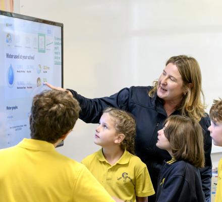 A classroom group gathers around a large interactive display showing school water-use data. One adult points to information on the screen while four primary school students in yellow school uniforms look on. The display includes graphs, water consumption figures and rainfall information as part of a water conservation or sustainability activity.