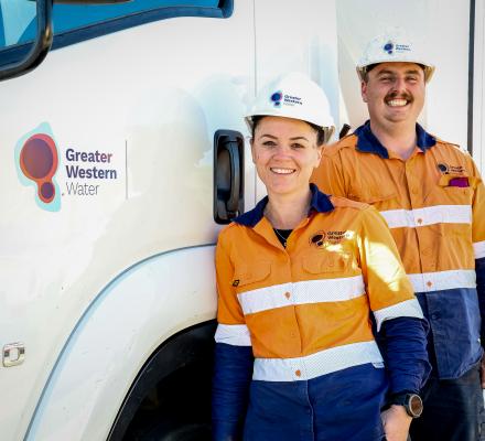 Two Greater Western Water field workers wearing high-visibility orange and navy uniforms and white safety helmets stand beside a white service truck displaying the Greater Western Water logo. The workers are positioned next to the truck door, with the vehicle filling most of the background in a bright outdoor setting.